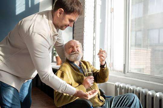 an elder man seated and holding a glass of water while another man stands behind him