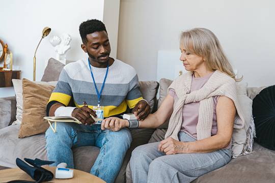 male carer checking a medical device on an elder woman's wrist while both are seated on a couch