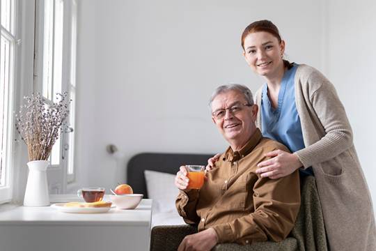 A female carer standing behind an elder man holding a glass of juice with breakfast on a table in front on him