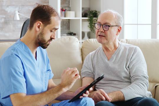 A male carer taking notes and discussing with an elder man while both are seated on a couch in the living room