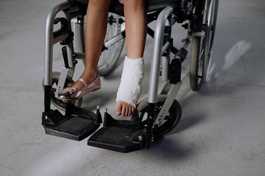 A woman sitting in a wheelchair, legs visible from the knees down, with one leg in an orthopedic cast.